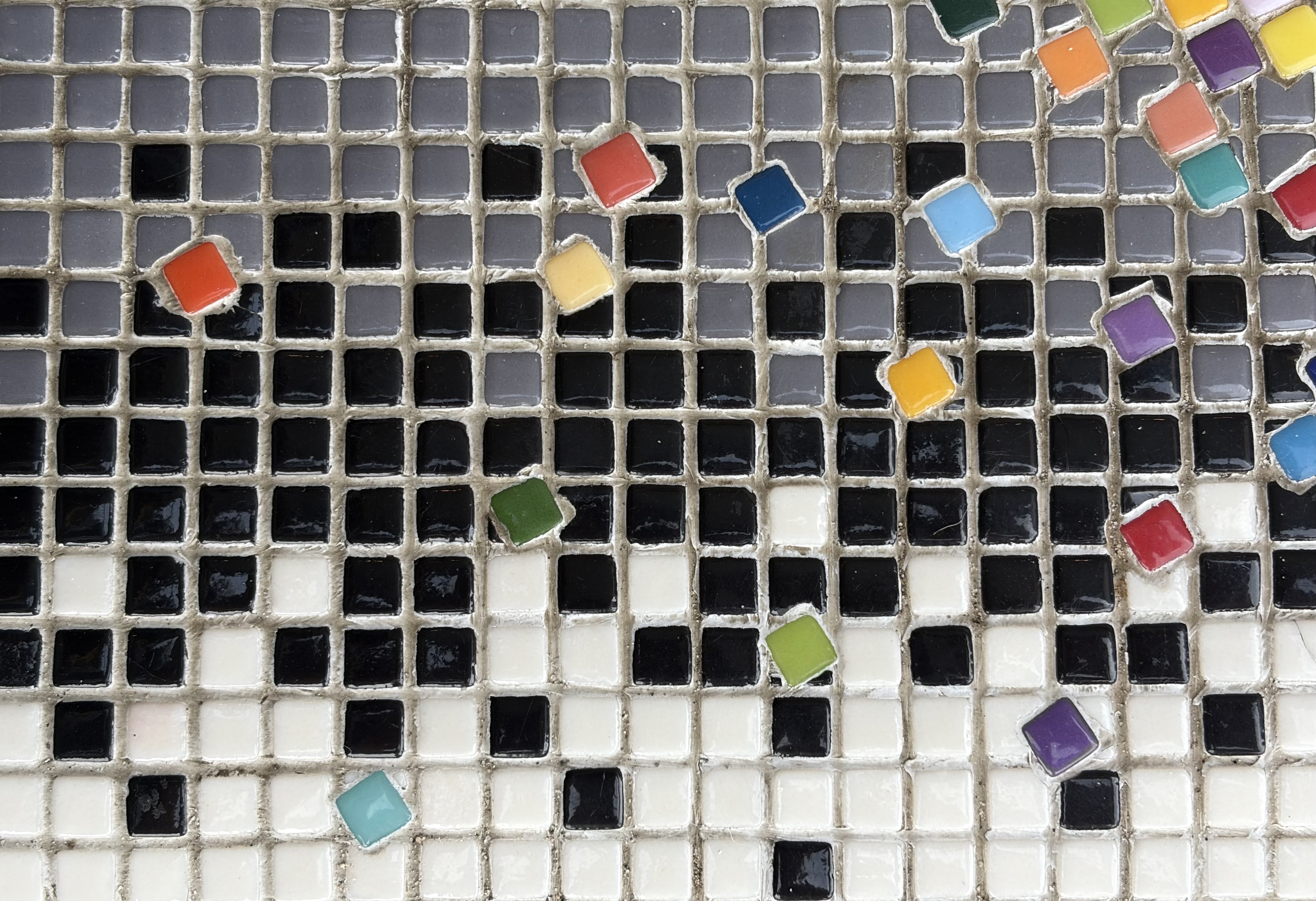 Mosaic detail of a black and white gradient tile grid with scattered rainbow-colored tesserae breaking through