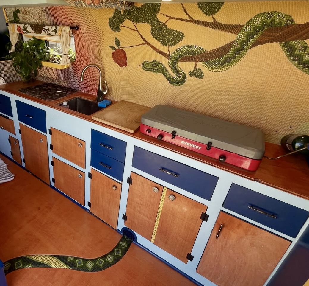Deception mosaic installed above a van kitchen counter, with cabinetry and a mosaic-tiled snake motif along the floor trim