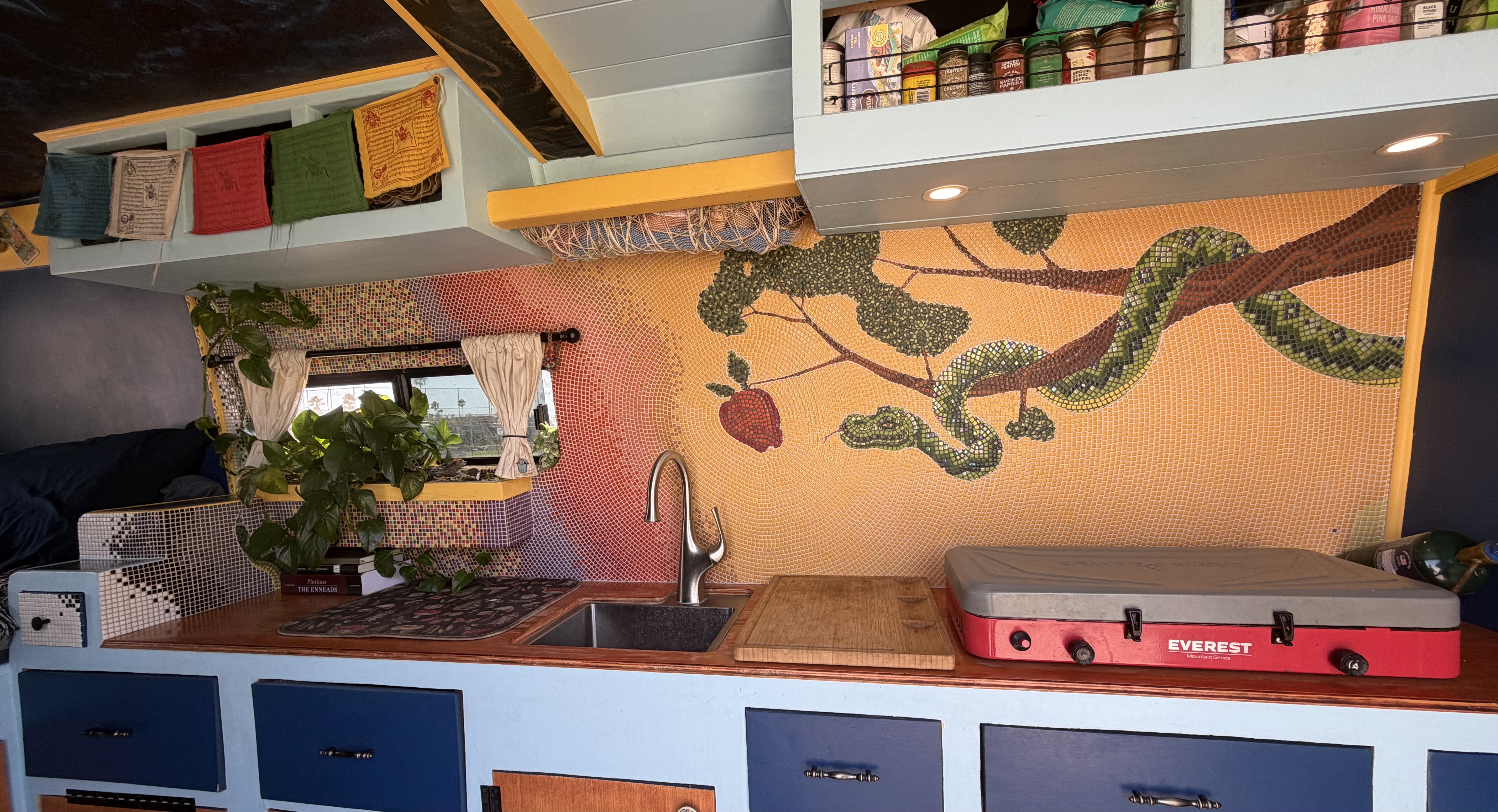 Full view of the van kitchen with the Deception mosaic, prayer flags, spice rack, and stovetop