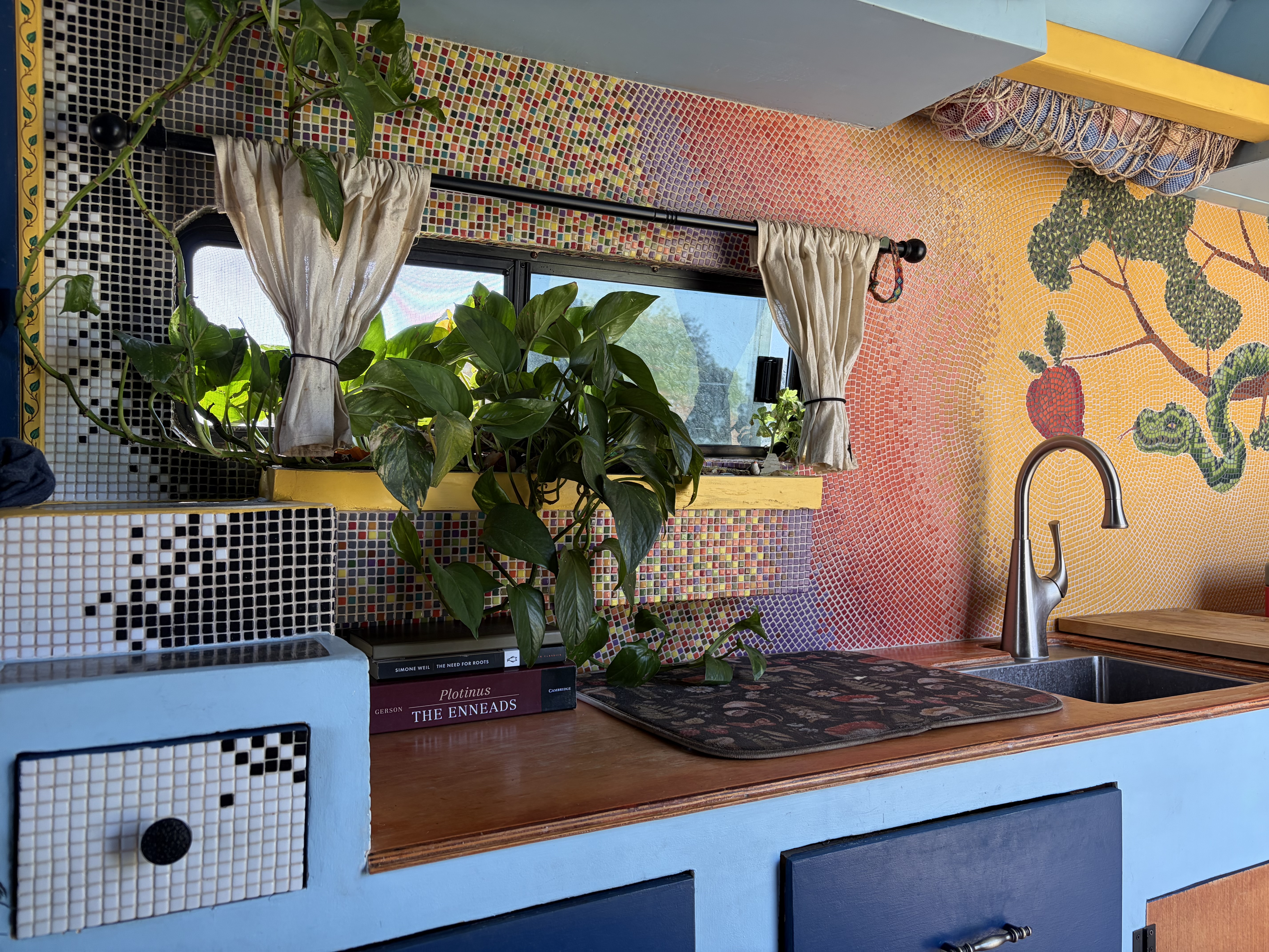 Deception mosaic surrounding a van kitchen window, with houseplants and a stack of philosophy books on the counter